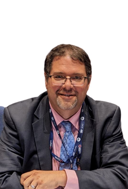 A man with short brown hair, glasses, and a beard is smiling. He is wearing a gray suit, pink shirt, and blue patterned tie, with lanyards around his neck. The background is white.