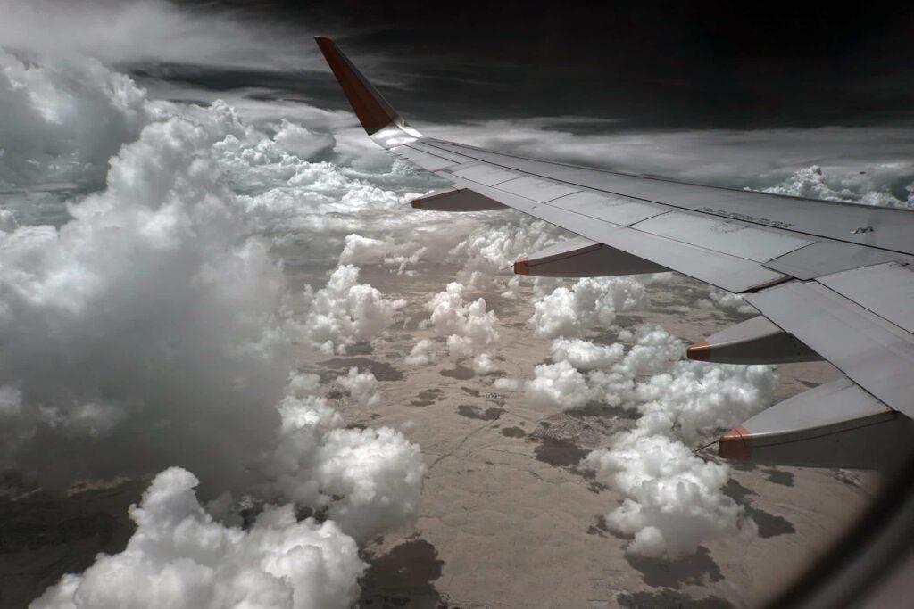 View from an airplane window showing the wing and winglet, flying above fluffy white clouds with a dramatic, dark sky and a landscape visible far below.