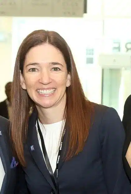 A woman with straight brown hair, wearing a navy blazer and white top, smiles at the camera in a brightly lit indoor setting. She has a black lanyard around her neck.