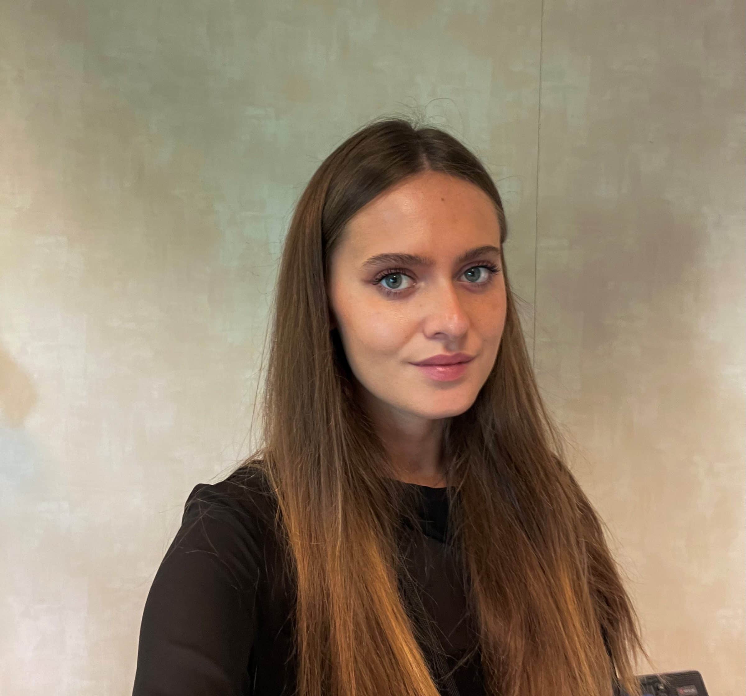 A young woman with long straight brown hair and blue eyes is looking at the camera, wearing a black top. She is standing indoors against a neutral beige wall.