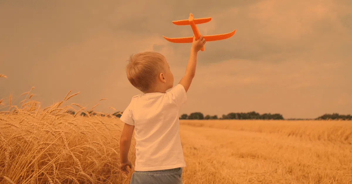 A young boy in a white t-shirt holds up an orange toy airplane while standing in a golden wheat field under a cloudy, orange-tinted sky.