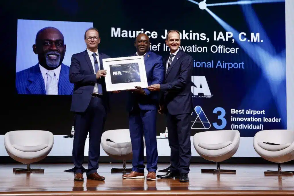Three men in suits stand on stage, smiling and holding a framed award. Behind them is a large screen displaying a portrait of one man and text recognizing Maurice Jenkins as an innovation leader at Miami International Airport.