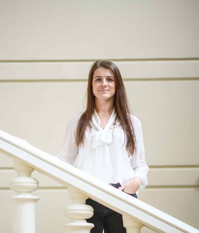 A young woman with long brown hair stands indoors beside a white staircase railing, wearing a white blouse and dark pants, with a neutral background featuring horizontal lines.