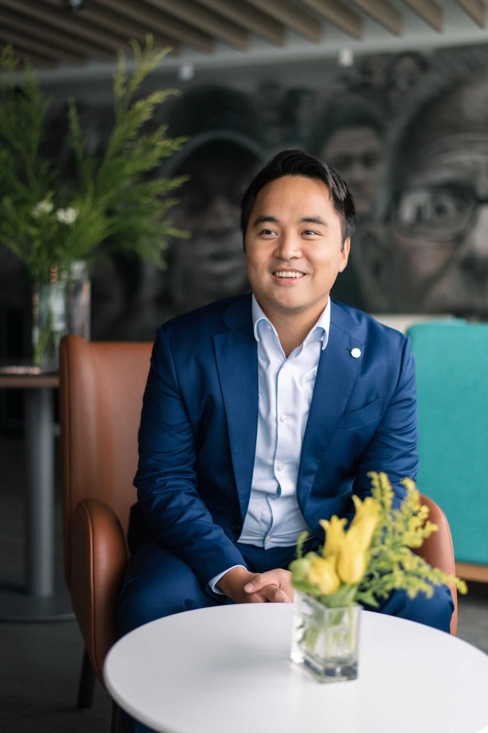 A man in a blue suit sits in a brown chair, smiling, with his hands resting on a white table holding a small glass vase of yellow flowers. The background includes green plants and a mural of faces.