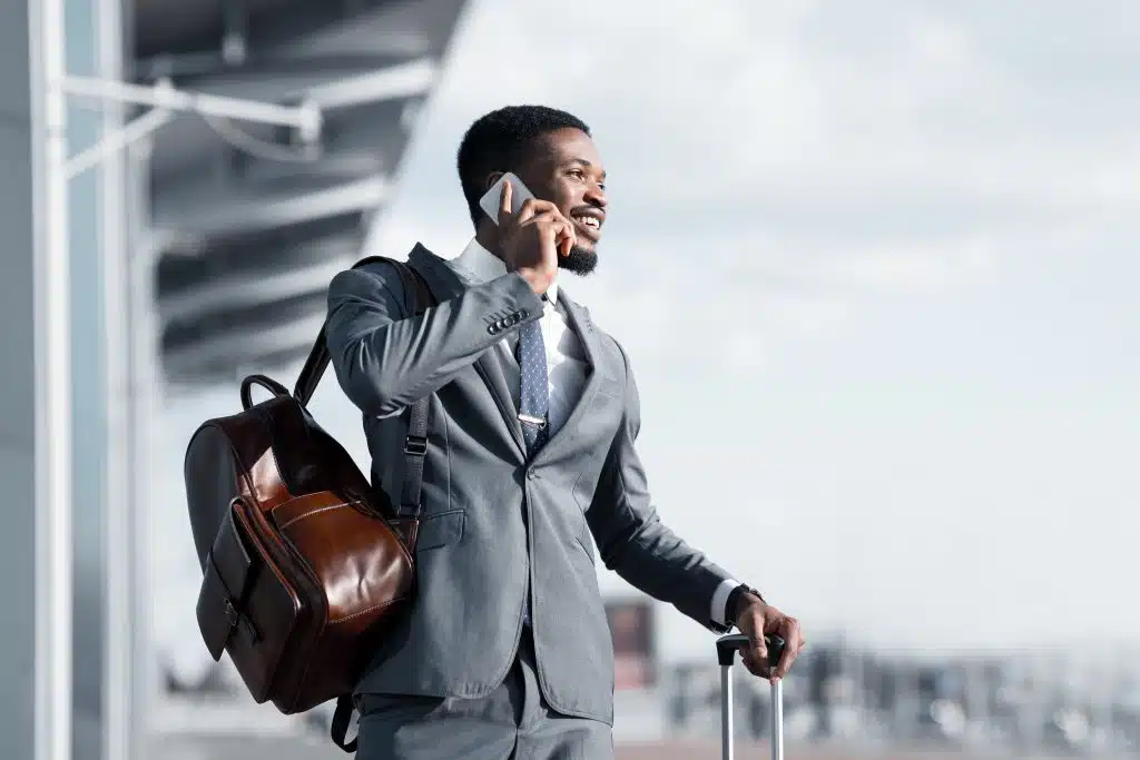A man in a gray suit stands outdoors, smiling while talking on his smartphone about an airport project management task. He carries a brown leather bag over his shoulder and holds the handle of a suitcase, suggesting he is traveling.