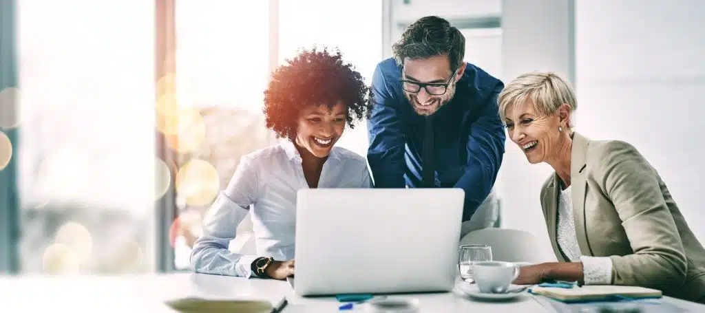 Three business professionals—two women and one man—smile and look at a laptop screen together in a bright office setting, appearing engaged in collaborative airport training.