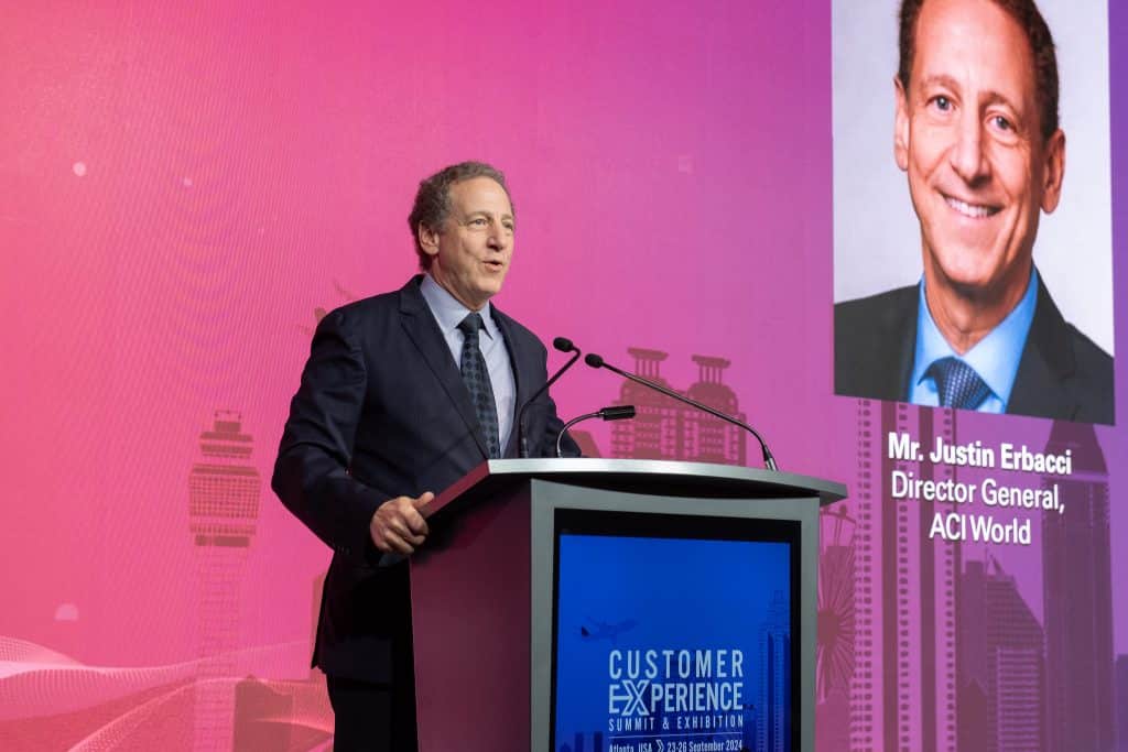 A man in a suit speaks at a podium during the Customer Experience Summit & Exhibition. Behind him, cityscape graphics and "Year of Transformation" are featured, along with a screen showing Mr. Justin Erbacci, Director General, ACI World.