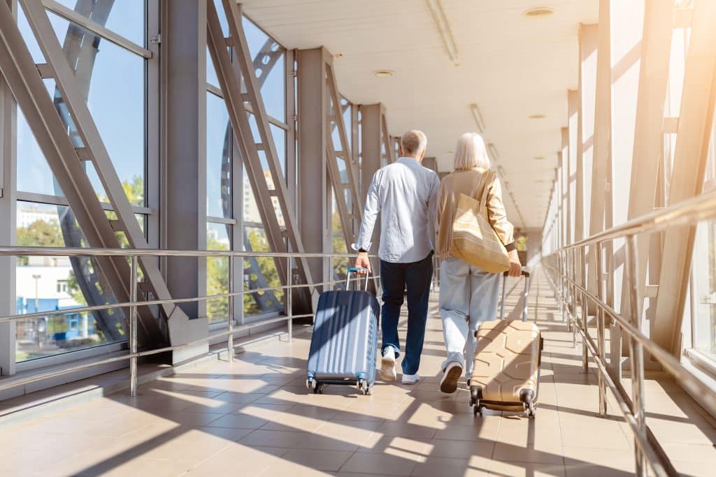 An older couple walks through an airport corridor, each pulling a rolling suitcase. Sunlight streams through large windows as they head toward their destination, passing signs for airport security along the way.