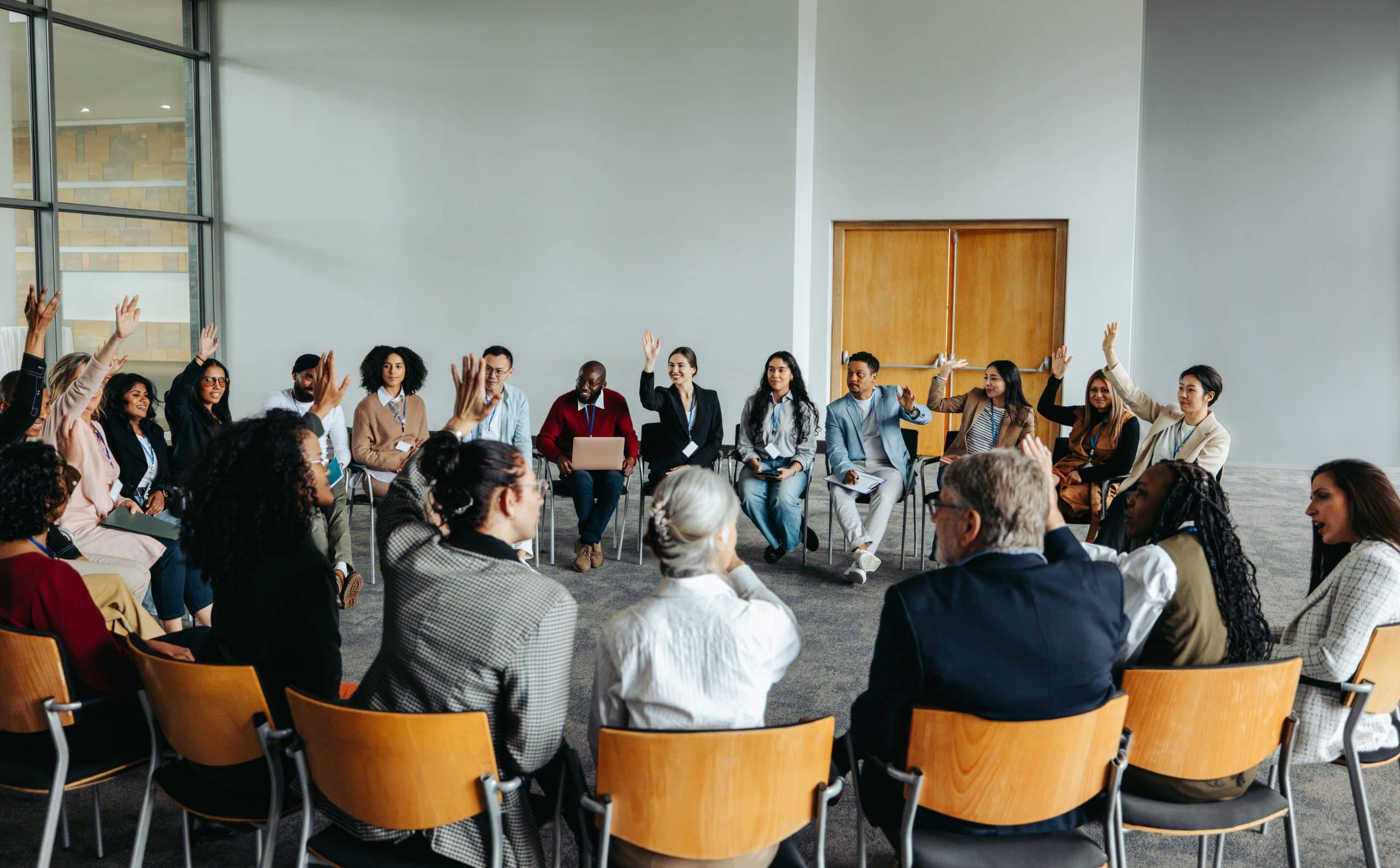 A diverse group of people sits in a circle indoors, engaging in a discussion about airport experience management. Several have their hands raised, suggesting they are volunteering ideas or responding to questions on improving passenger journeys.