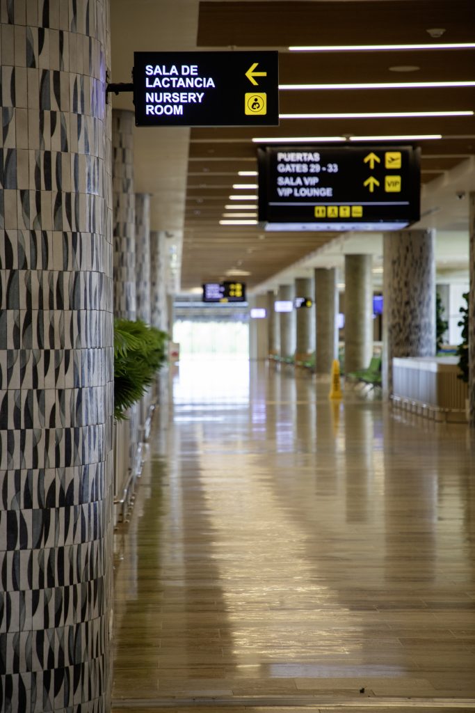 A modern airport hallway with patterned walls, plants, and overhead signs in Spanish and English—including a nod to Women's Day—directing to the nursery room, gates 29–33, and the VIP lounge. The corridor is empty and well-lit.