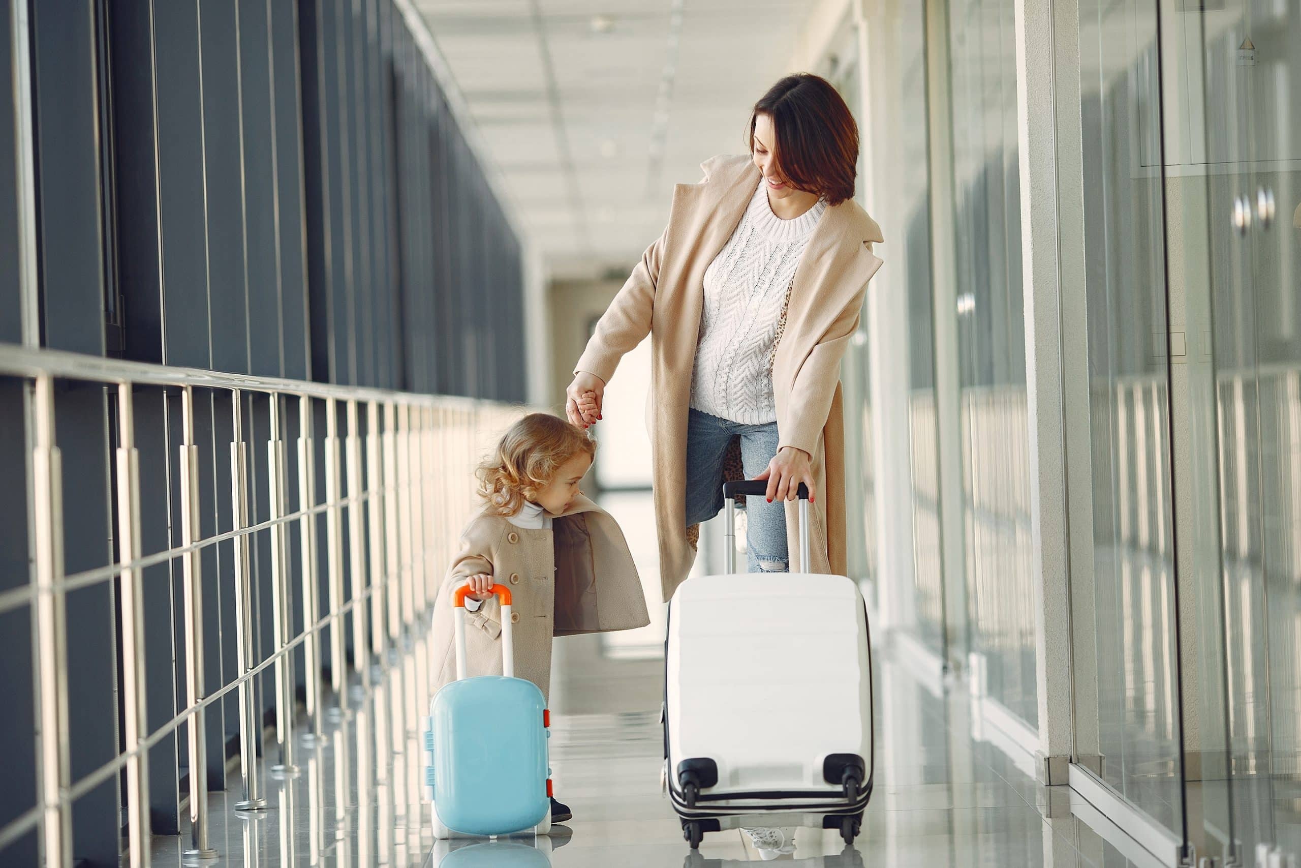 A woman and a young child, both in light-colored coats, walk hand in hand down an airport hallway on Women's Day. The woman pulls a white suitcase while the child follows with a small blue suitcase.